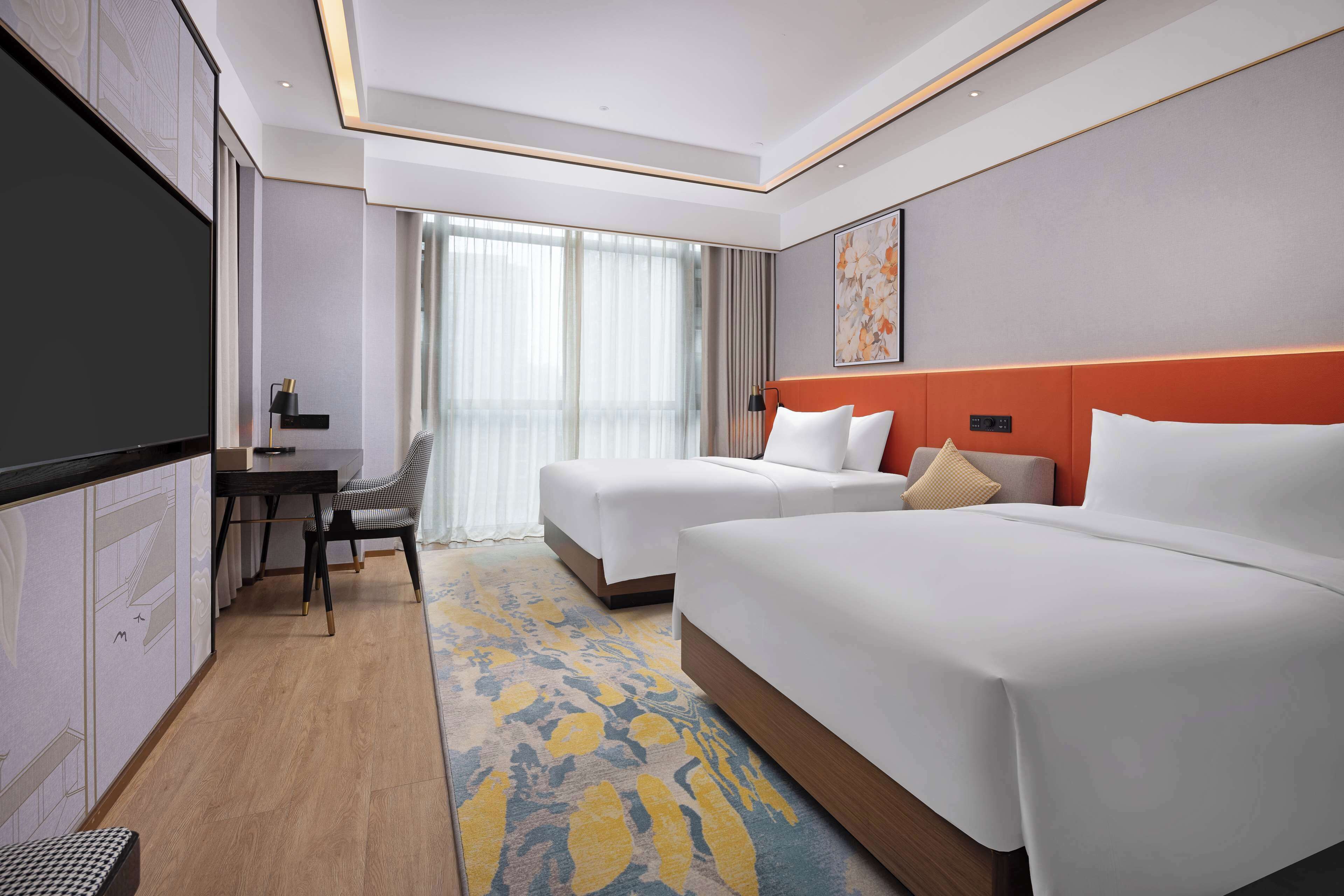 A hotel room with two beds, a desk with a chair, a flat-screen TV, and a large window with curtains.