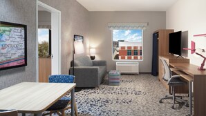 Une chambre d’hôtel de taille modeste, avec un canapé gris, une petite table, un téléviseur et un bureau équipé d’un ordinateur.