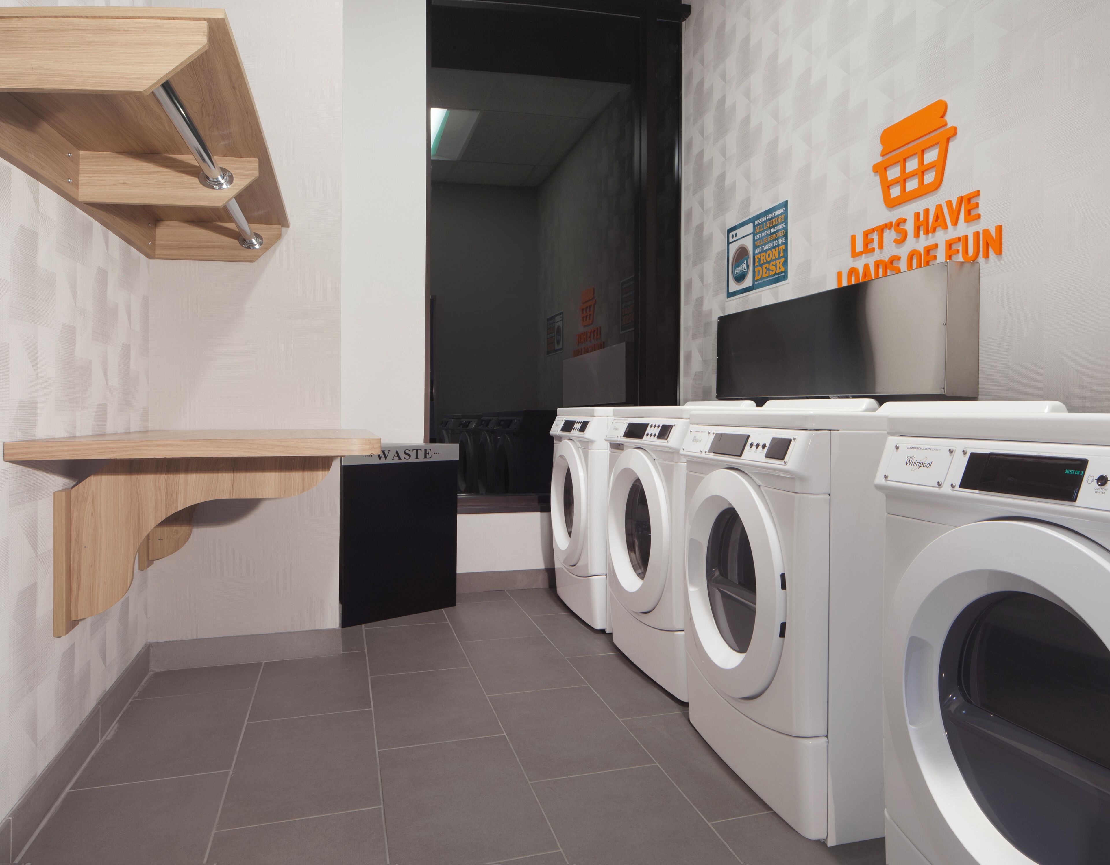 A laundry area with a wooden shelf, a waste bin, and multiple front-loading washing machines.