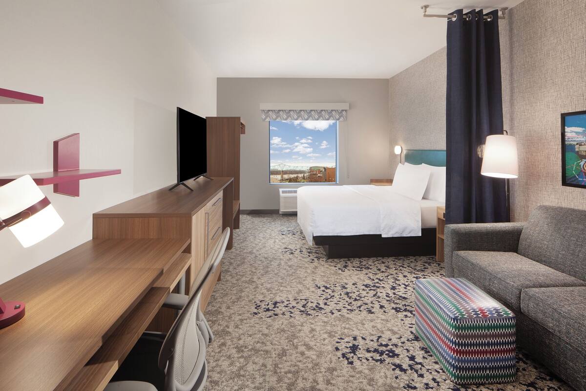 Habitación de hotel con cama, escritorio con computadora, silla, sofá, ventana con vistas y lámpara.