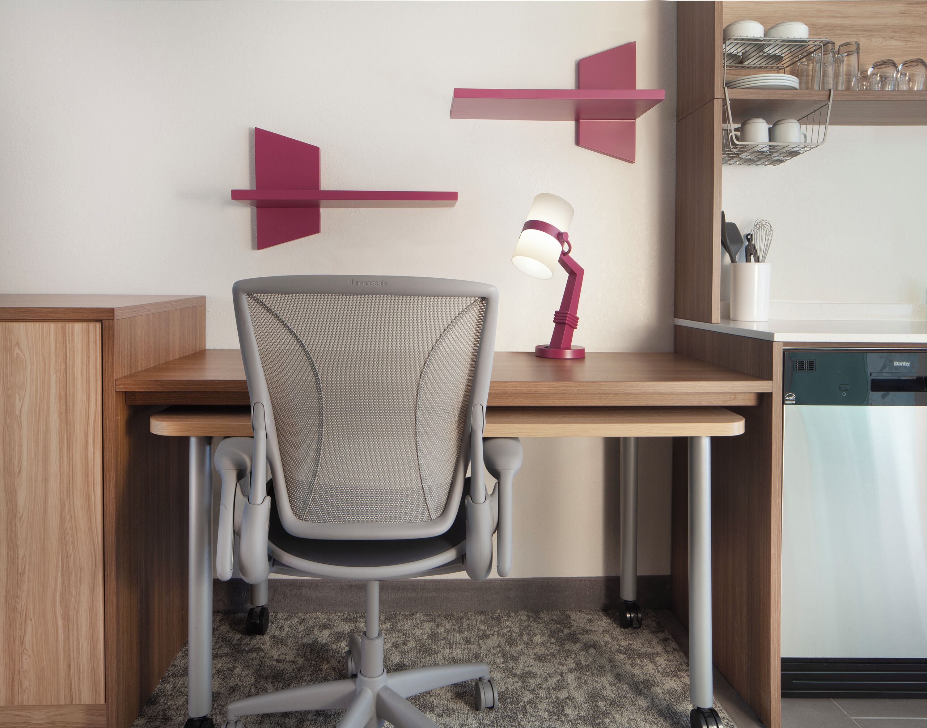 A modern office chair with a mesh backrest, a desk with a wooden finish, and a red desk lamp.