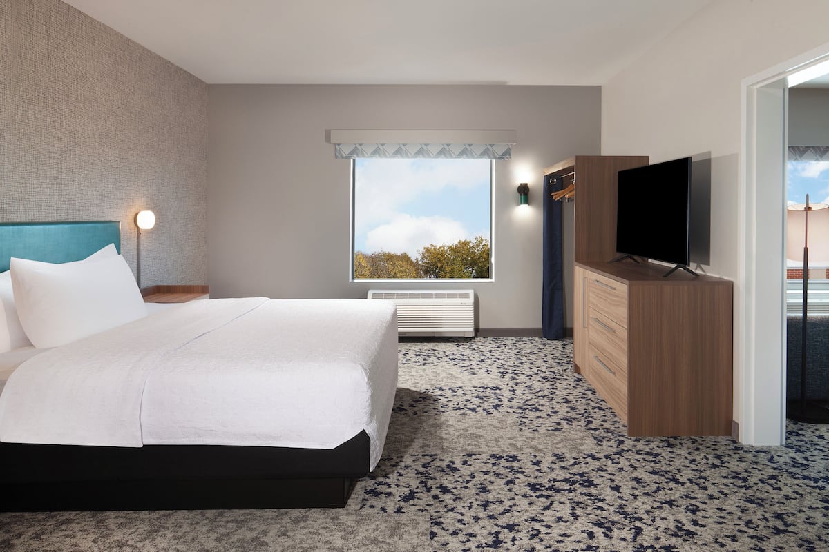 Habitación de hotel moderna con una cama grande, un televisor de pantalla plana, un aparador de madera y un ventanal con vistas a árboles.