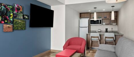 A modern living room with a grey sofa, a red armchair, and a red coffee table. A flat-screen TV is mounted on the wall, and there are framed pictures of coffee plants.