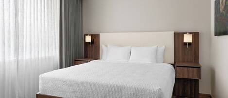 A modern hotel room with a large bed, wooden headboard, two bedside tables with lamps, and a view of the city through sheer curtains.