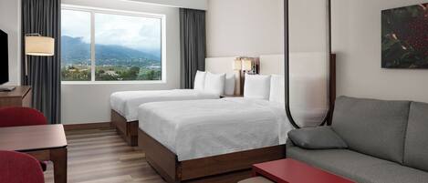 Quarto de hotel com duas camas, um sofá, uma escrivaninha e uma grande janela com vista para as montanhas.