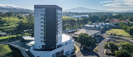 A modern hotel with a prominent tower labeled 'HOMEWOOD SUITES by Hilton,' surrounded by lush greenery and a clear sky.