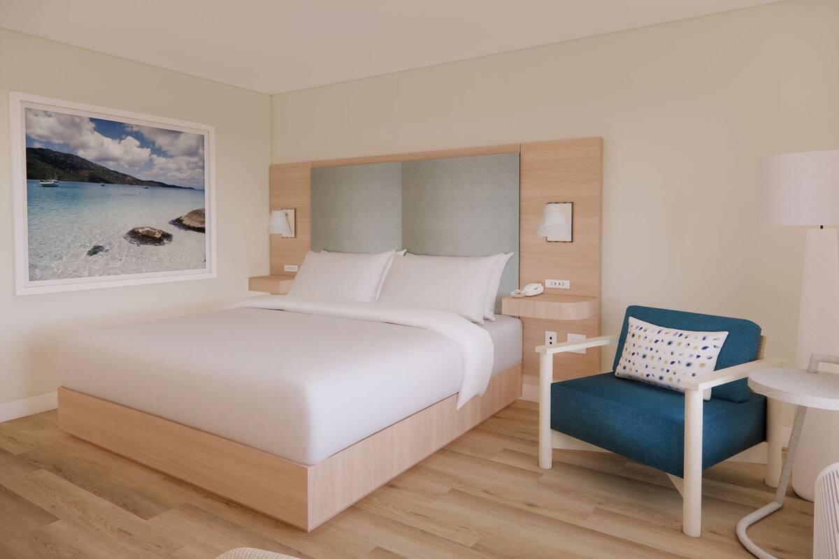 A hotel room with a bed, a chair, a small table, and a wall-mounted lamp.