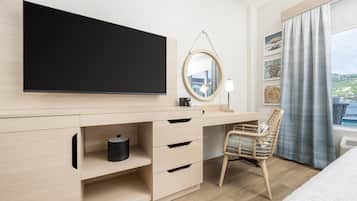 A modern hotel room with a large flat-screen TV, a wooden desk with drawers, a wicker chair, and a large window with a view of the outdoors.