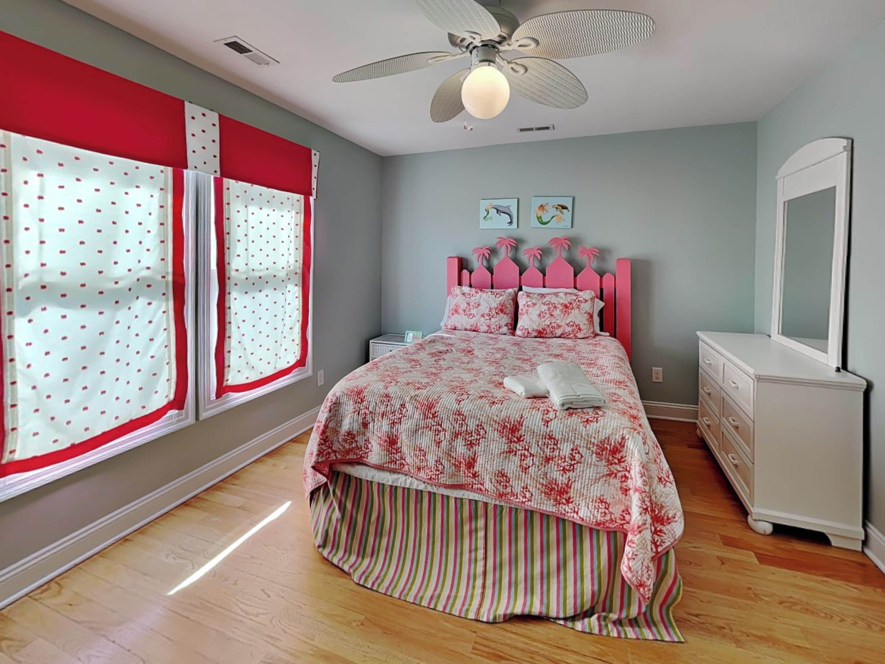 Un dormitorio con cama, cómoda, ventilador de techo y ventana con cortinas.