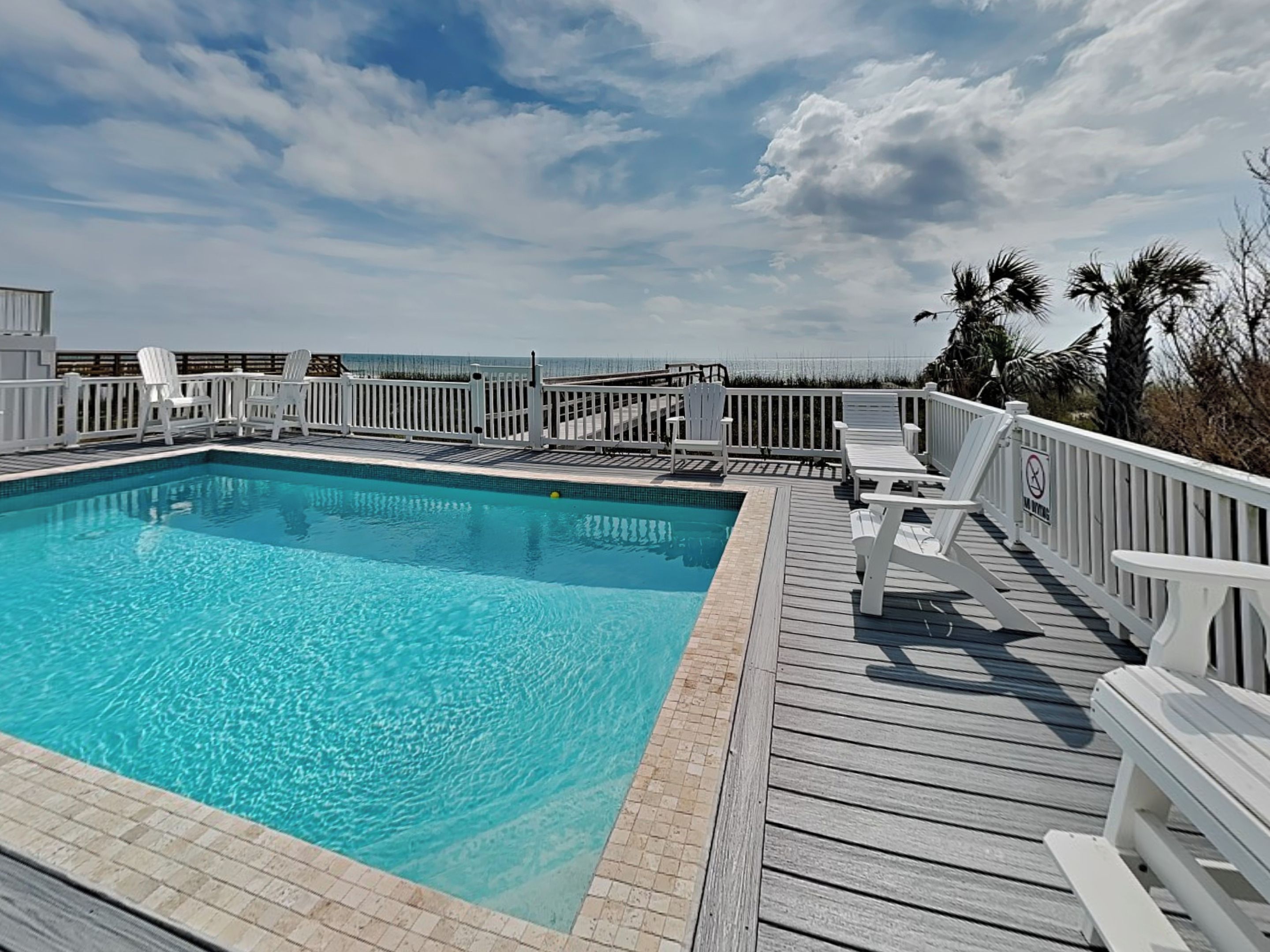 A rectangular pool with a wooden deck and white railings, surrounded by beach chairs and palm trees.