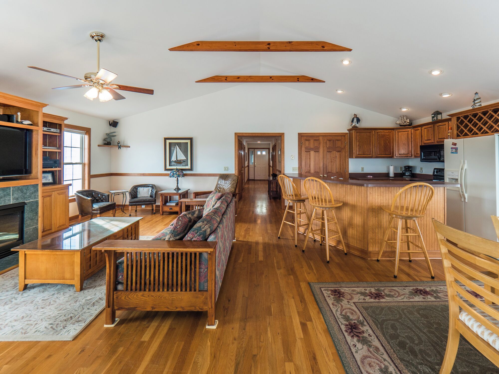 A spacious living area with a sofa, chairs, and a dining table. There is a kitchen with wooden cabinets and a refrigerator. A ceiling fan is visible, and the room has wooden beams on the ceiling.