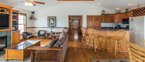 A spacious living area with a sofa, chairs, and a dining table. There is a kitchen with wooden cabinets and a refrigerator. A ceiling fan is visible, and the room has wooden beams on the ceiling.