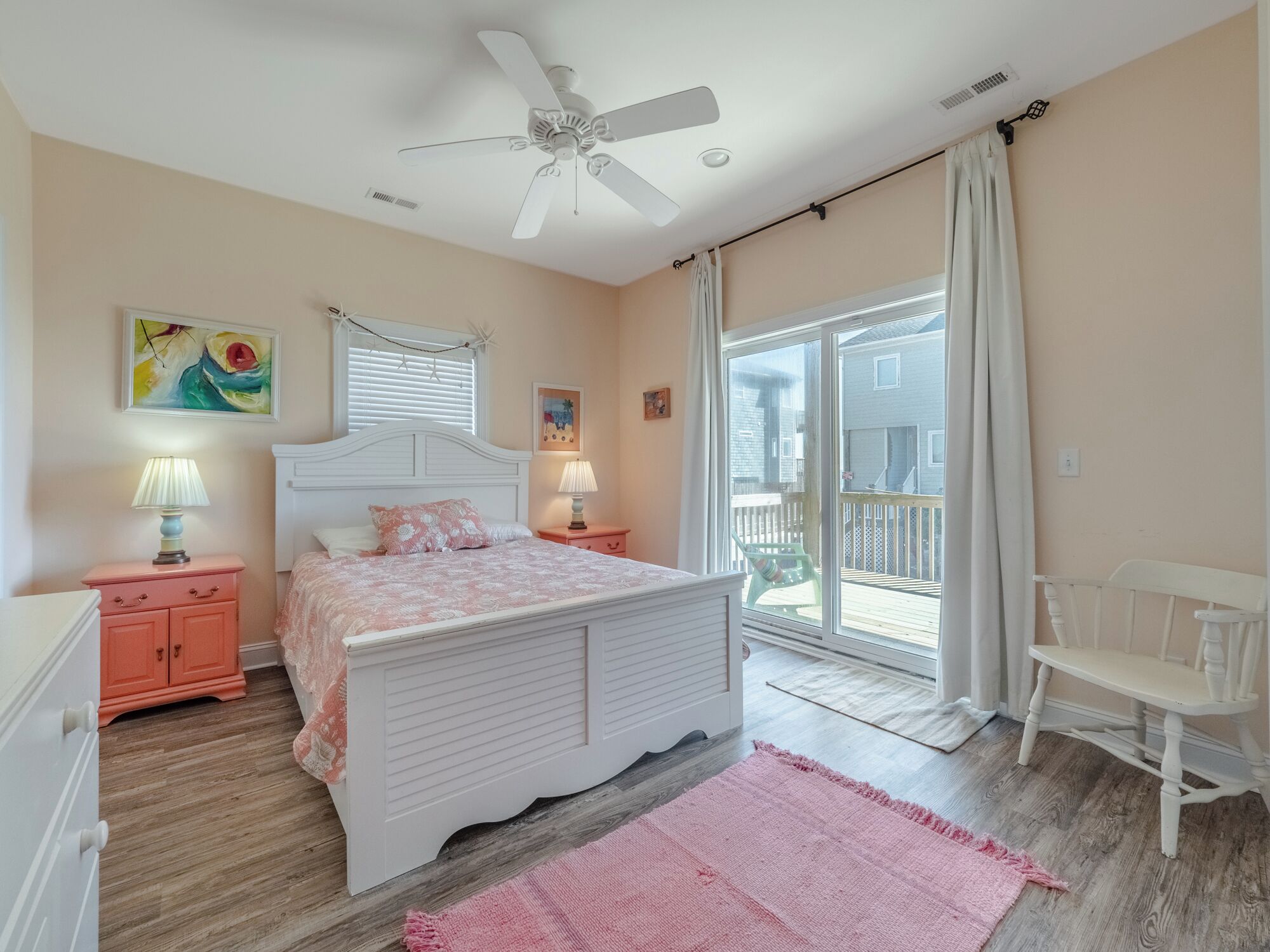 A bedroom with a bed, a ceiling fan, a window with blinds, a nightstand, a lamp, a chair, and a view of the outside.