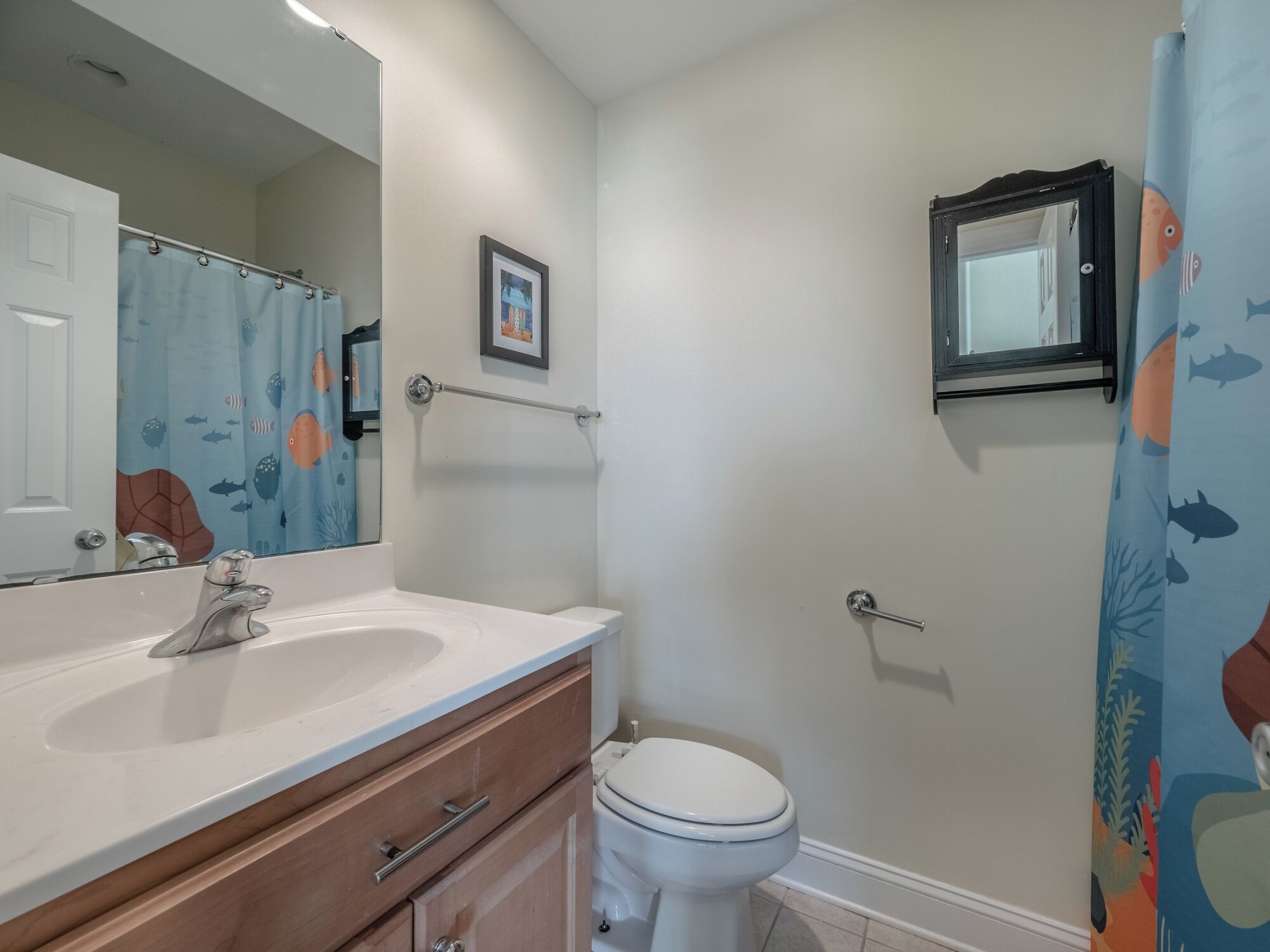 A bathroom with a white sink, a mirror, a framed picture on the wall, and a shower curtain with a fish pattern.