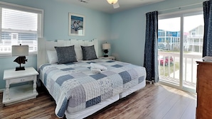 A bedroom with a bed, a nightstand, a ceiling fan, a window with blinds, and a view of buildings outside. - Shoo Fly by Carolina Retreats (Kure Beach)