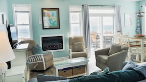 A living room with a television, two armchairs, a glass coffee table, and a view of the ocean through large windows. - Sunflower Beach House by Topsail Realty (Holly Ridge)