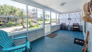 A covered porch with blue flooring, two wicker chairs, a small table, and a view of a residential area with trees and buildings. - Ankers Away by Topsail Realty (Holly Ridge)