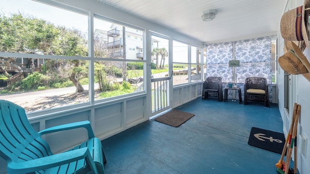 A covered porch with blue flooring, two wicker chairs, a small table, and a view of a residential area with trees and buildings. - Ankers Away by Topsail Realty (Holly Ridge)