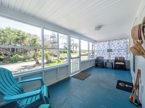 A covered porch with blue flooring, two wicker chairs, a small table, and a view of a residential area with trees and buildings. - Ankers Away by Topsail Realty (Holly Ridge)