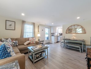 A spacious living room with a sofa, coffee table, and a dining area with a table and chairs. There is a kitchen in the background with white cabinets and a large mirror above the sink. - Ankers Away by Topsail Realty (Holly Ridge)