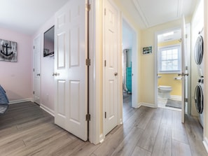 Una habitación con piso de madera, puertas blancas, un cuadro en la pared y vista a un baño con inodoro y lavadora.