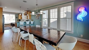 A spacious dining area with a large wooden table surrounded by white chairs, a kitchen in the background, and a neon squid sign on the wall. - A Shore Cure by Carolina Retreats (Kure Beach)