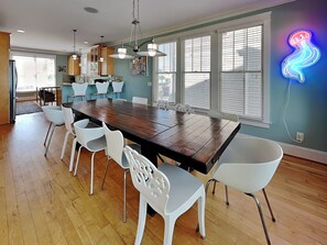 A spacious dining area with a large wooden table surrounded by white chairs, a kitchen in the background, and a neon squid sign on the wall.
