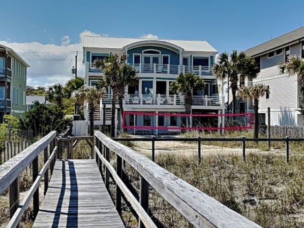 A beachfront property with a wooden boardwalk leading to a multi-story building with balconies and a pool area.