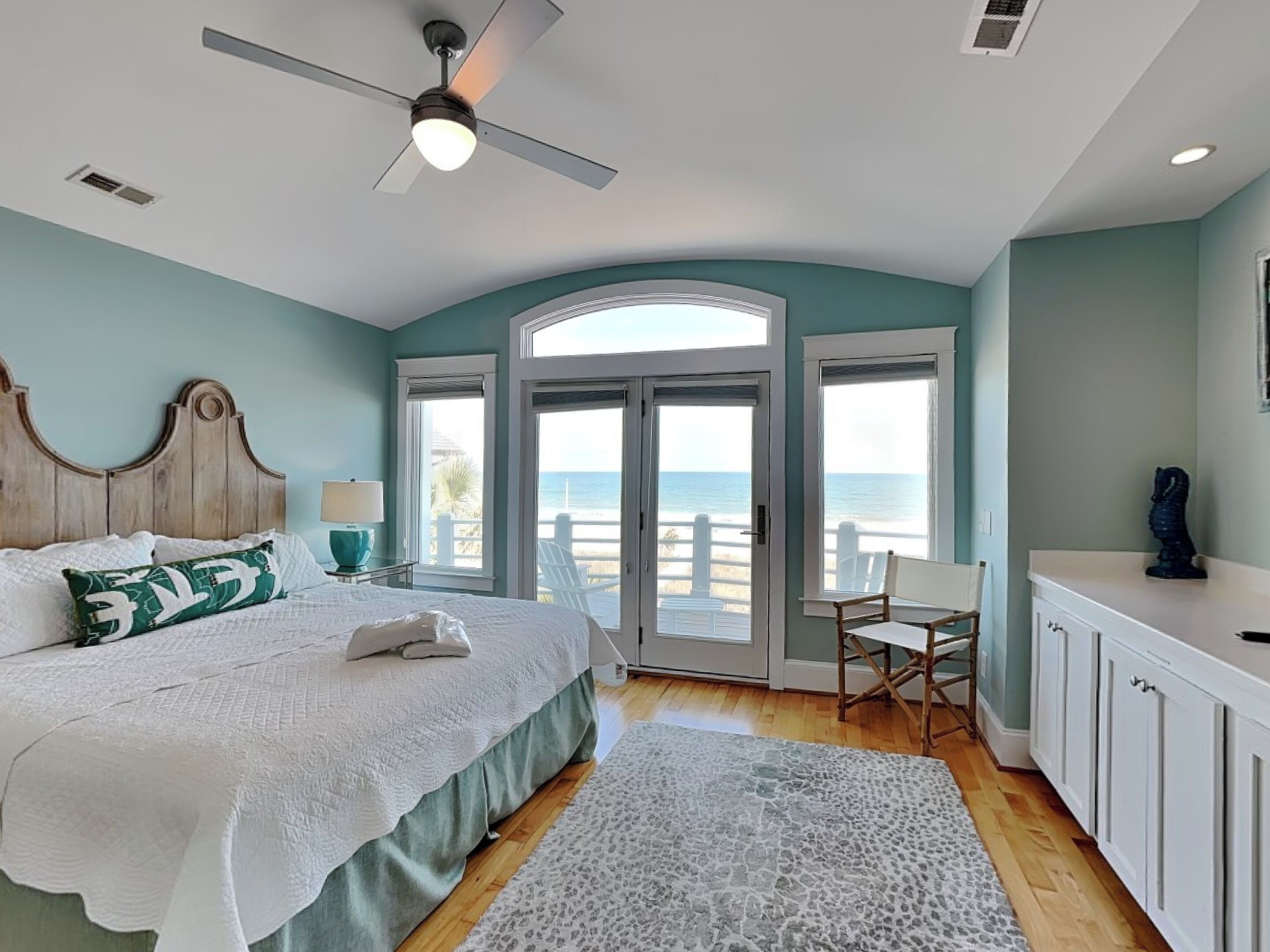 A bedroom with a bed, a ceiling fan, a view of the beach, a chair, and a desk.