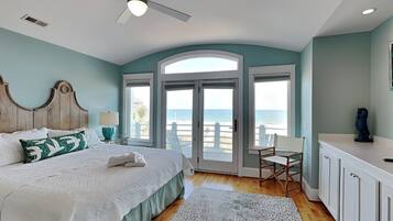 A bedroom with a bed, a ceiling fan, a view of the beach, a chair, and a desk.