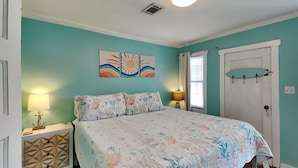 A bedroom with a bed, two nightstands, a door with a coat rack, and a wall with a sunburst painting. - CB Sunshine Cottage by Carolina Retreats (Carolina Beach)