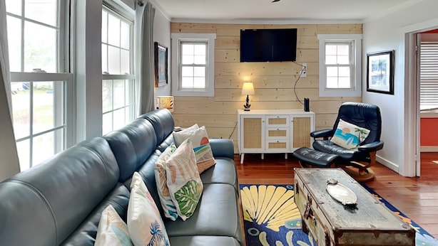 A living room with a leather sofa, a wooden cabinet, a chair, and a ceiling fan. - CB Sunshine Cottage by Carolina Retreats (Carolina Beach)