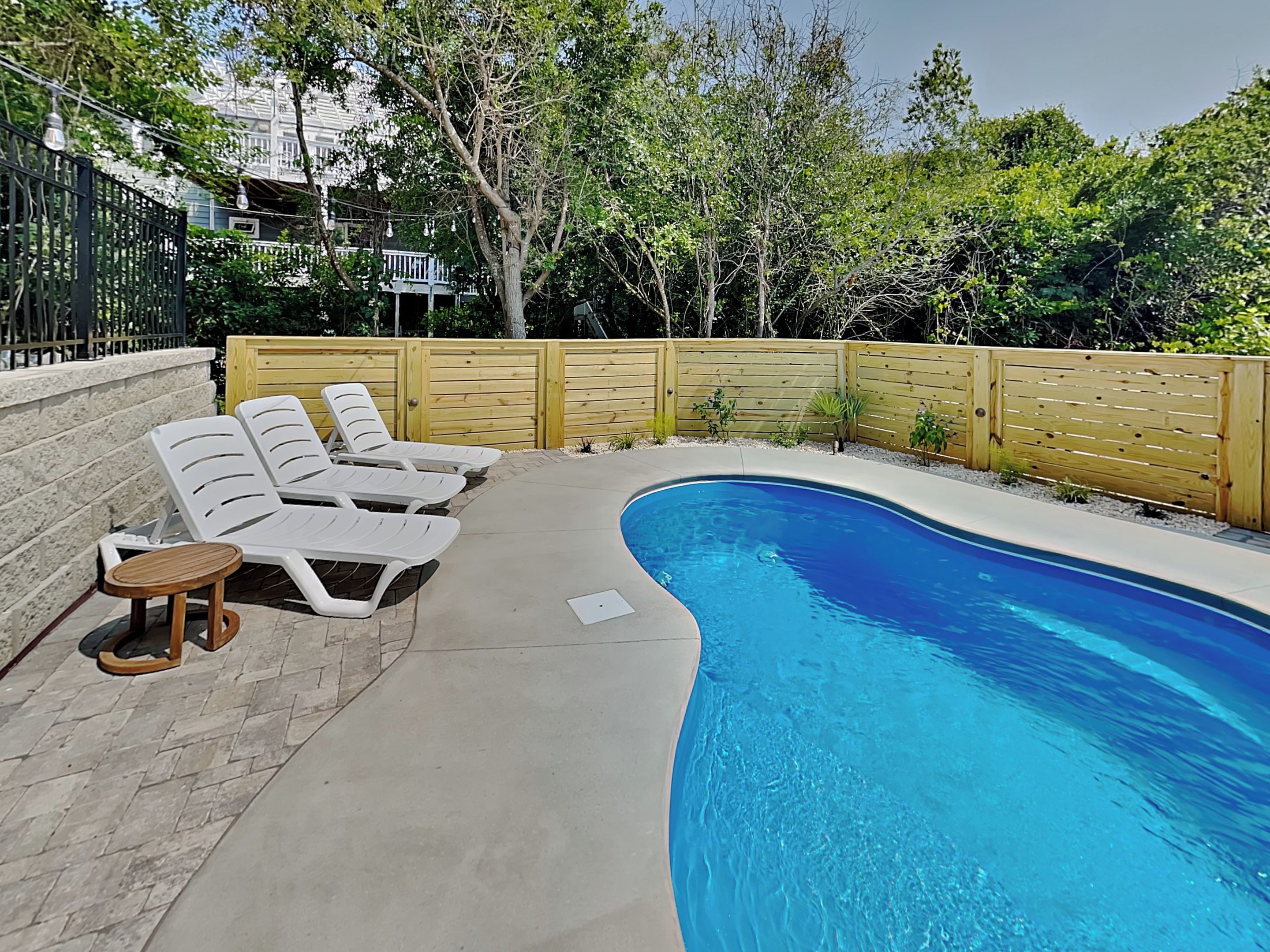 A swimming pool with lounge chairs and a small table, surrounded by a wooden fence and trees.