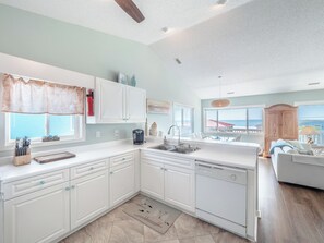 A modern kitchen with white cabinets, a dishwasher, and a large window overlooking the ocean. - Blue Haven by Topsail Realty (Holly Ridge)