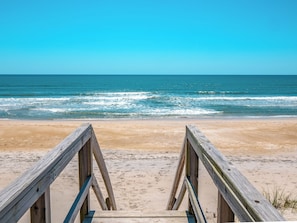 Une promenade en bois qui mène à une plage où l’eau est d’un bleu limpide et le rivage est sablonneux.