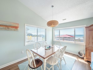 A dining area with a table and chairs, large windows with blinds, a hanging light fixture, and a view of buildings and the ocean. - Blue Haven by Topsail Realty (Holly Ridge)