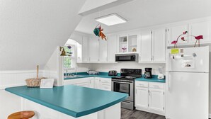 A kitchen with white cabinets, a teal countertop, and a central island with stools. - Just Beachy Spartanburg by Carolina Retreats (Carolina Beach)