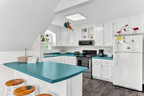 A kitchen with white cabinets, a teal countertop, and a central island with stools. - Just Beachy Spartanburg by Carolina Retreats (Carolina Beach)