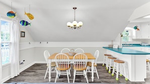 A dining area with a wooden table and white chairs, a kitchen with a blue countertop, and a ceiling with hanging fish decorations. - Just Beachy Spartanburg by Carolina Retreats (Carolina Beach)