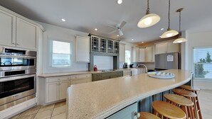 A modern kitchen with stainless steel appliances, a granite countertop, and a bar area with stools. - Over The Moon by Carolina Retreats (Kure Beach)