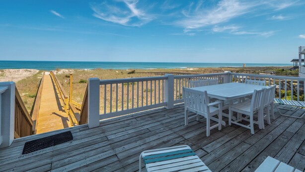 A wooden deck with white chairs and a table overlooking a beach. - Over The Moon by Carolina Retreats (Kure Beach)