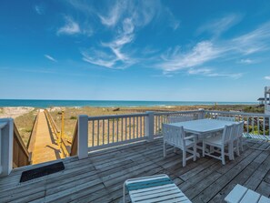 A wooden deck with white chairs and a table overlooking a beach. - Over The Moon by Carolina Retreats (Kure Beach)