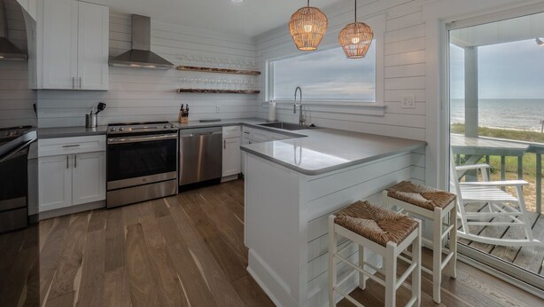 A modern kitchen with white cabinetry, stainless steel appliances, and a large island with bar stools. - Summer Place by Carolina Retreats (Kure Beach)