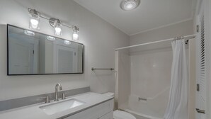 A modern bathroom with a white sink, vanity, and toilet. There is a shower with a curtain and a mirror above the sink. - Summer Place by Carolina Retreats (Kure Beach)