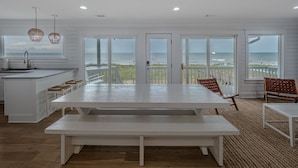 A spacious kitchen with a large island, white cabinetry, and a view of the ocean through large windows. - Summer Place by Carolina Retreats (Kure Beach)