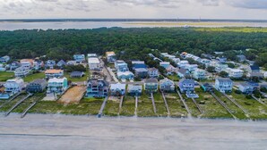An aerial view of a coastal residential area with houses aligned along the beachfront, a sandy beach, and a forested background. - Summer Place by Carolina Retreats (Kure Beach)