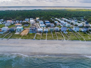 Vue aérienne d’une zone résidentielle côtière, avec des maisons alignées le long du bord de mer, une plage de sable et un paysage boisé en arrière-plan.