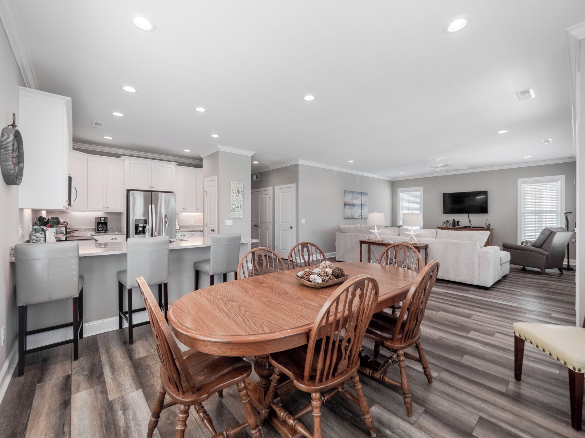A spacious living area with a wooden dining table, grey upholstered chairs, a grey sofa, and a flat-screen TV mounted on the wall.