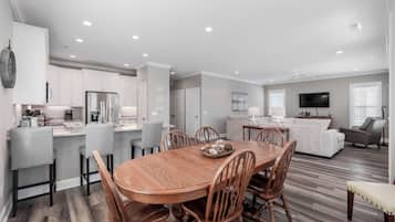 A spacious living area with a wooden dining table, grey upholstered chairs, a grey sofa, and a flat-screen TV mounted on the wall.