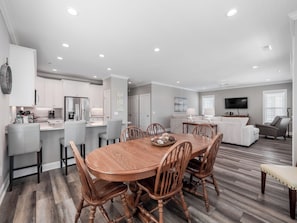 A spacious living area with a wooden dining table, grey upholstered chairs, a grey sofa, and a flat-screen TV mounted on the wall. - A Kure Cure by Topsail Realty (Kure Beach)